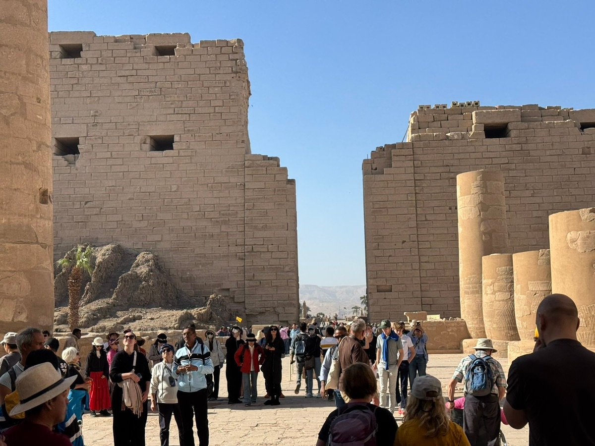 View of scattered ruins and dry shrubbery at Karnak, with parts of the temple in the background.
