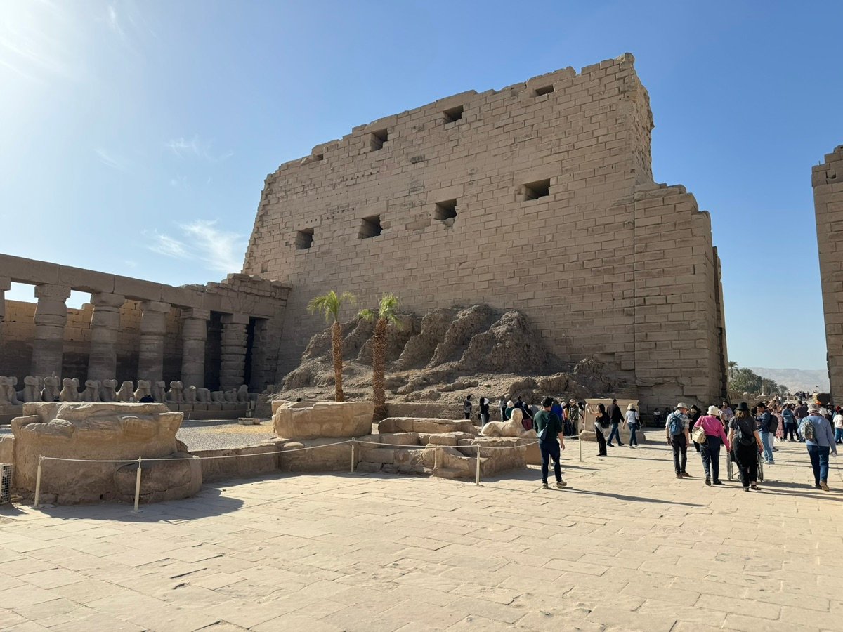 A dusty path through ancient ruins with palm trees and a large stone structure in the background.