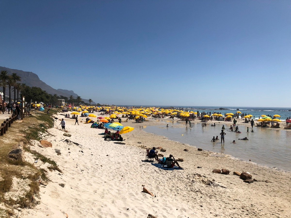 People relaxing on the grassy area next to Camps Bay Beach, with a blue tent in the foreground